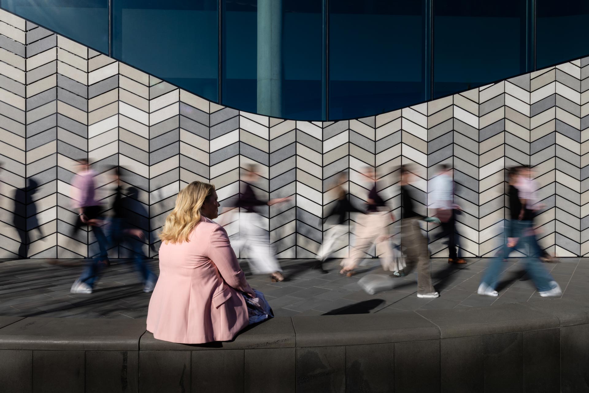 Camille seated still while a crowd blurs past a chevron-tiled wall