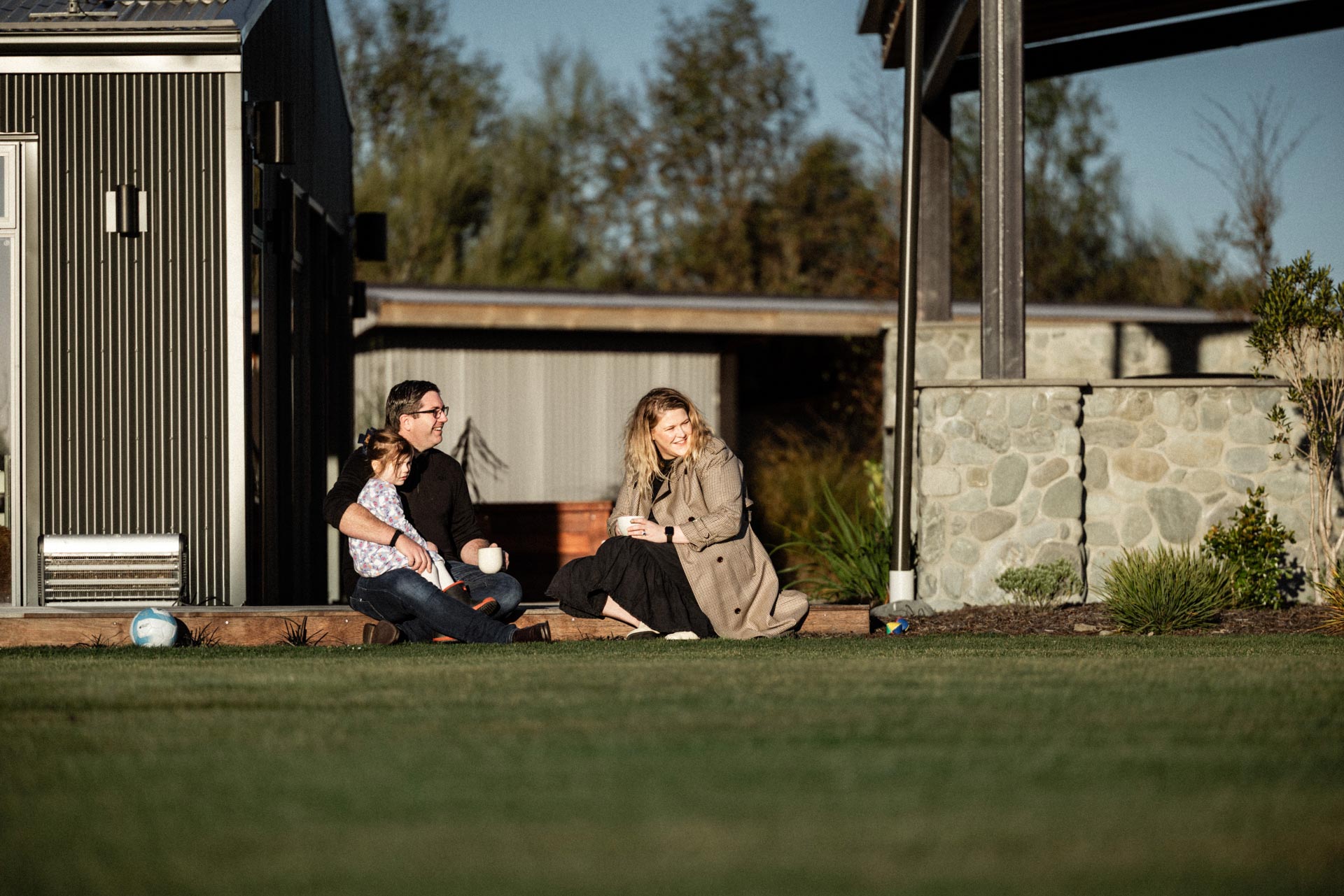Camille, Chris and their daughter sitting on the lawn with a cuppa at Whakahā